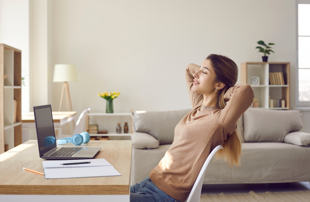 Woman in living room leaning back in chair with eyes closed at a desk with an open laptop and notepad