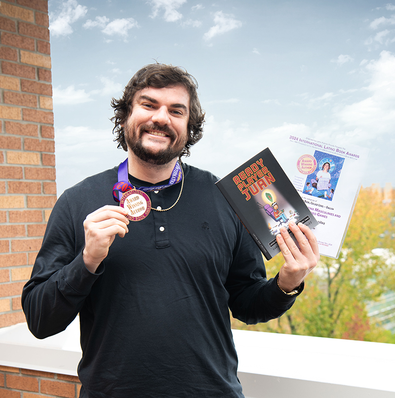 Photo of Carlos Kelly, Ph.D on the balcony of the Norman J. Radow College of Humanities and Social Sciences holding a medal for the 2024 International Latino Book Awards, and a copy of his winning book 