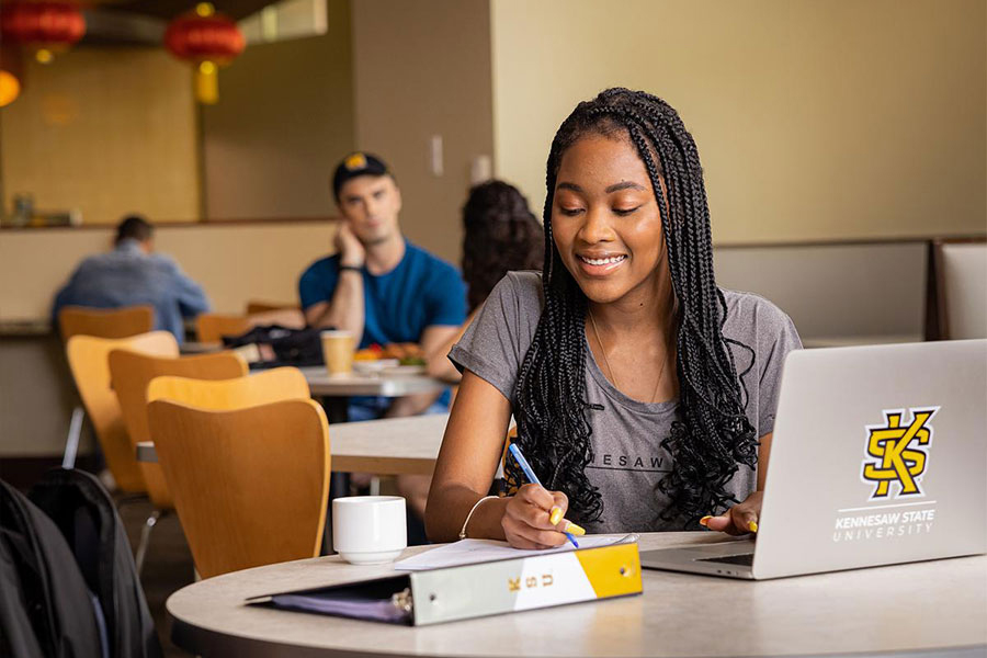 Student in student lounge jotting notes with laptop open